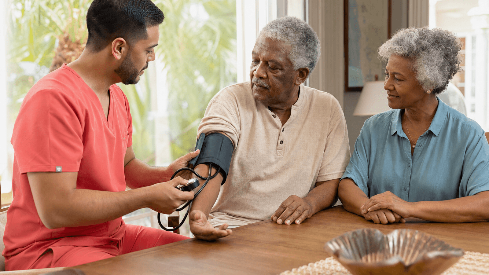 Hospice nurse in plain coral scrubs checking a patient's blood pressure during a home visit