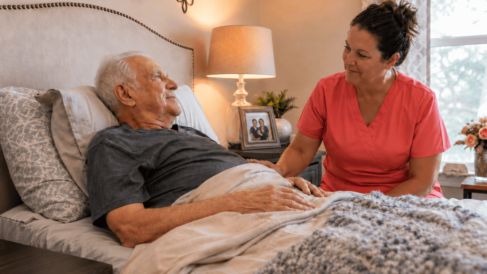 Hospice nurse in coral scrubs comforting an older patient at his bedside