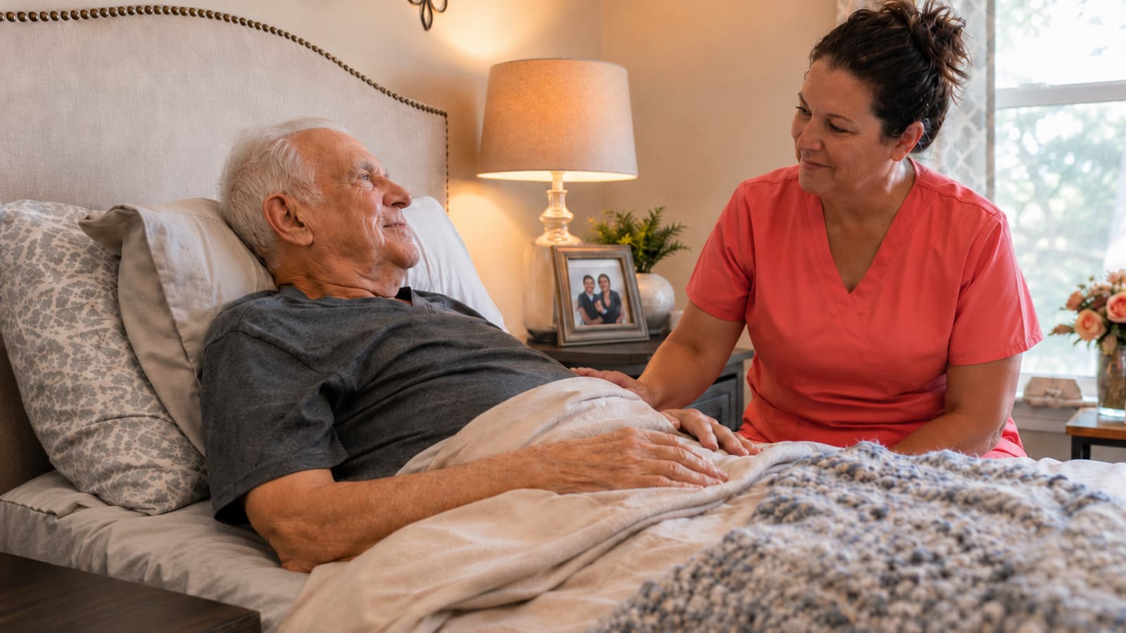 Hospice nurse in coral scrubs comforting an older patient at his bedside